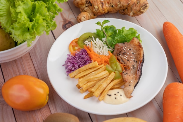 Filete de pescado con papas fritas, kiwi, lechuga, zanahorias, tomates y repollo en un plato blanco.