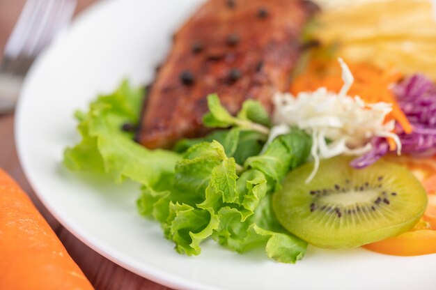 Filete de pescado con papas fritas, kiwi, lechuga, zanahorias, tomates y repollo en un plato blanco.