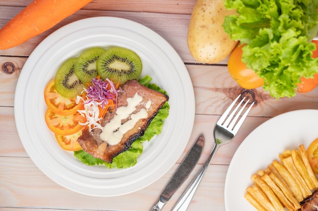Filete de pescado con papas fritas, kiwi, lechuga, zanahorias, tomates y repollo en un plato blanco.