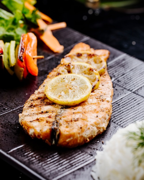 Filete de pescado a la parrilla con palitos de limón y vegetales en un tablero de filete negro.