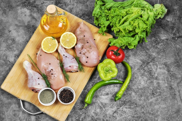Filete de pollo crudo y piernas en placa de madera con verduras frescas, especias y vaso de aceite.