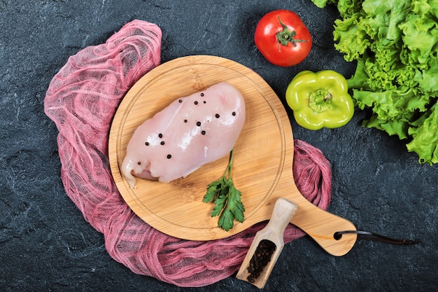 Filete de pollo crudo sobre tabla de madera con verduras y mantel.