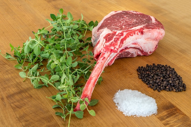 Filete de Tomahawk angus crudo sobre una tabla de madera con sal, pimienta, orégano y un cuchillo