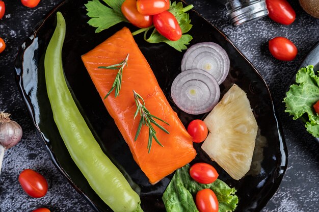 Filetes de salmón crudo, pimienta, kiwi, piña y romero en un plato y piso de cemento negro.