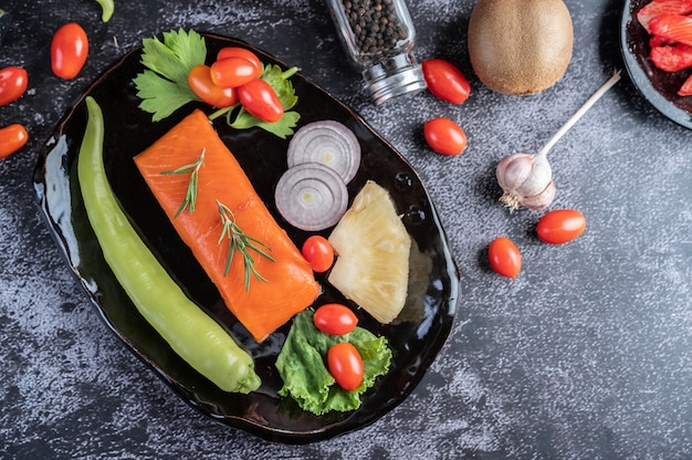 Foto gratuita filetes de salmón crudo, pimienta, kiwi, piña y romero en un plato y piso de cemento negro.