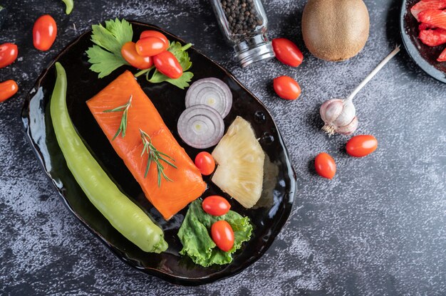 Filetes de salmón crudo, pimienta, kiwi, piña y romero en un plato y piso de cemento negro.