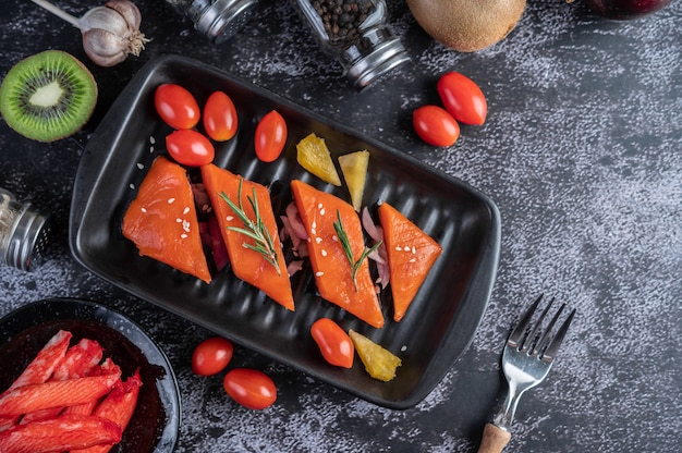 Filetes de salmón crudo, pimienta, kiwi, piña y romero en un plato y piso de cemento negro.