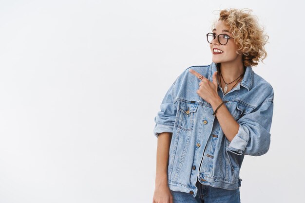 Filmación en interiores de una agradable chica de ensueño con estilo con el pelo corto y rubio en gafas y chaqueta de mezclilla