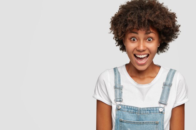 Filmación en interiores de alegre mujer de piel oscura vestida con peto, se ríe positivamente