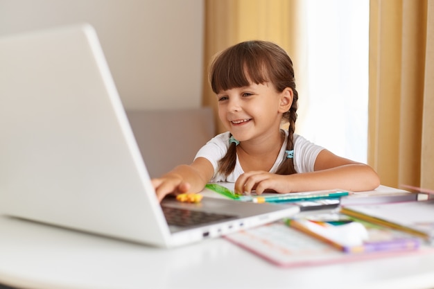 Filmación en interiores de un alumno de pelo oscuro positivo feliz posando en casa, mirando la computadora portátil con una sonrisa encantadora, educación a distancia, con lecciones en línea.