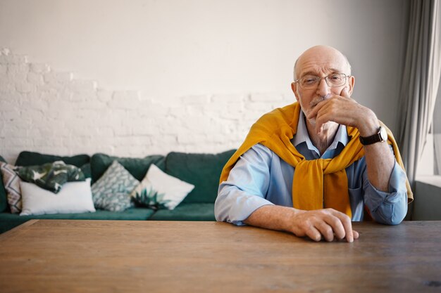 Filmación en interiores de apuesto anciano maduro con ojos sabios sentado en un escritorio de madera con sofá con expresión pensativa, tocando la cara. Personas, estilo de vida y edad