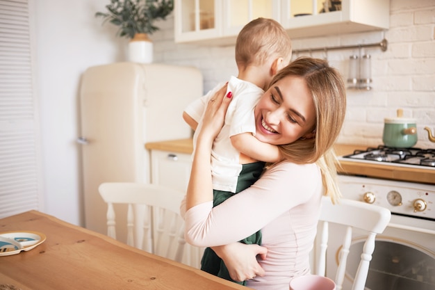 Foto gratuita filmación en interiores de una atractiva joven madre rubia que pasa un buen rato en casa abrazando a un niño pequeño sentado en la mesa de comedor en la acogedora cocina, sonriendo, disfrutando de felices momentos dulces de su maternidad