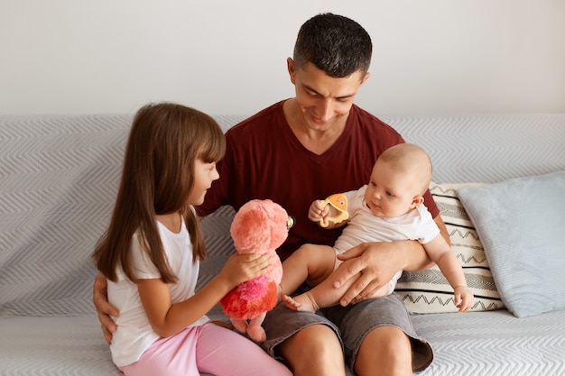 Filmación en interiores de un atractivo hombre morena vestido con camiseta marrón pasar tiempo con sus hijos, sentados en el sofá en la sala de luz, jugando juntos, expresando felicidad.