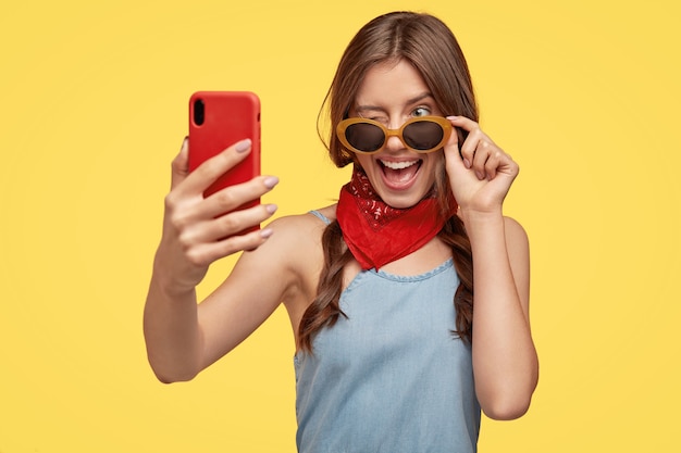 Filmación en interiores de complacida joven morena con gafas de sol posando contra la pared amarilla