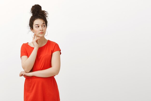 Filmación en interiores de curiosa mujer seria con cabello rizado peinado en moño, vistiendo un vestido rojo