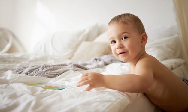 Filmación en interiores de un encantador niño europeo de un año con mejillas regordetas de pie al borde de la cama, tratando de trepar, extendiendo la mano para tomar el libro. Concepto de hora de dormir, sueño y cuidado de niños.