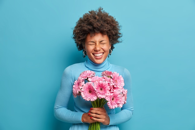 Filmación en interiores de encantadora joven de pelo rizado tiene una ocasión especial para recibir un regalo, tiene un bonito ramo de flores, adora las gerberas rosas