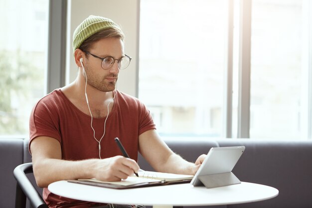 Filmación en interiores de un estudiante universitario serio con gafas y sombrero, escribe notas desde la tableta