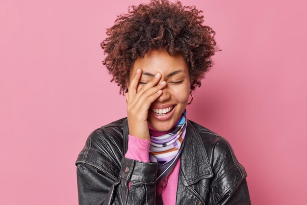 Filmación en interiores de feliz joven de pelo rizado mantiene los ojos cerrados con la mano en la cara sonríe suavemente viste chaqueta de cuero aislada sobre fundamento de estudio rosa. Concepto de emociones positivas y sentimientos sinceros