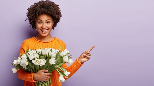 Filmación en interiores de feliz modelo de mujer joven con corte de pelo afro, apunta con el dedo índice