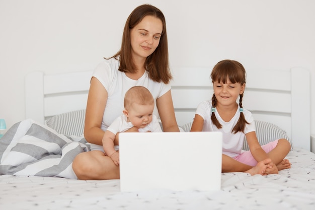 Filmación en interiores de feliz mujer positiva con cabello oscuro sentada en la cama con sus hijos, con dos niñas, tratando de trabajar desde perfeccionar, freelance y crianza de los hijos,