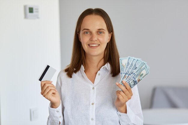 Filmación en interiores de feliz mujer rica positiva de pie en la sala de luz contra la pared blanca, sosteniendo billetes de dólar y tarjeta de crédito en las manos, mirando a cámara, vistiendo camisa.