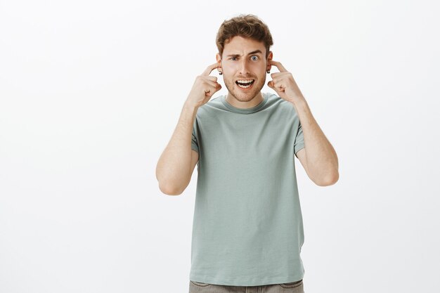 Filmación en interiores de gracioso novio caucásico en camiseta casual, haciendo muecas, cubriendo la audición con los dedos índices en los oídos