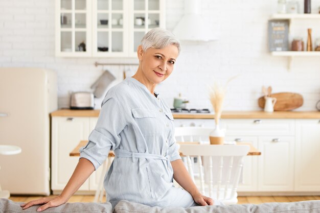 Filmación en interiores de hermosa mujer madura de pelo gris con elegante camisa azul posando contra el acogedor interior de la cocina, manteniendo las manos en la parte posterior del sofá. Elegante dama jubilada relajante en casa