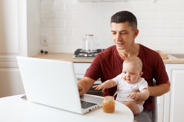 Filmación en interiores de un hombre autónomo guapo sonriente con camiseta burdeos, posando en la cocina blanca, sentado frente a la computadora portátil con el bebé en las manos, escribiendo en el cuaderno.