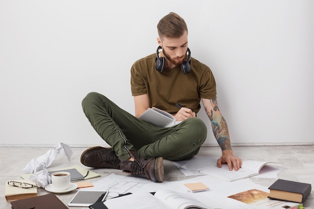 Filmación en interiores de hombre hipster con peinado moderno, berad grueso y brazos tatuados, mira atentamente el libro