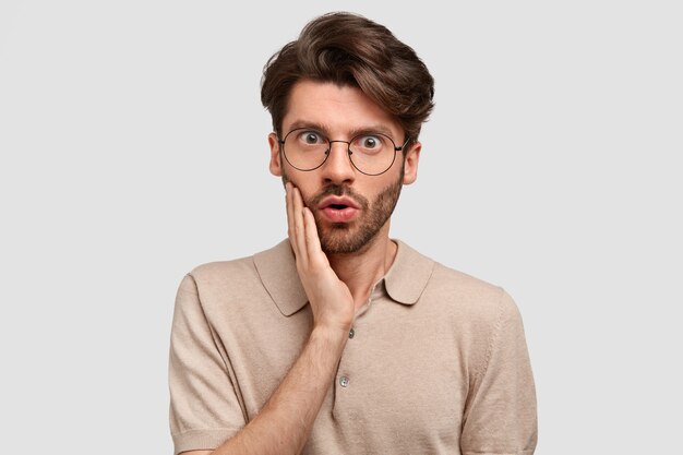 Filmación en interiores de un hombre serio barbudo con expresión asustada, toca la mejilla, tiene una barba oscura, vestido con ropa informal, está de pie contra la pared blanca