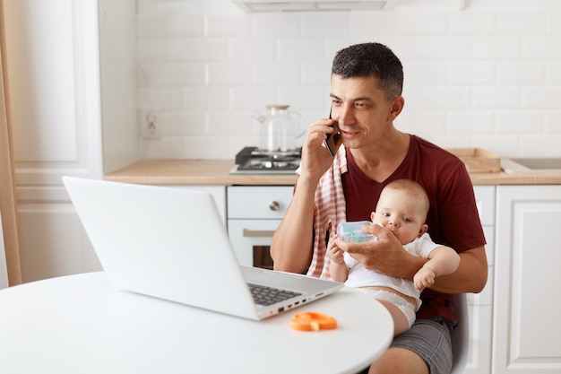 Filmación en interiores de un hombre vestido con una camiseta informal color burdeos con una toalla en el hombro, cuidando al bebé y trabajando en línea desde casa, hablando por teléfono y dándole agua a su hijo.