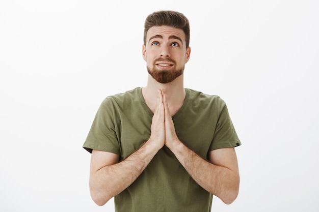 Filmación en interiores de un lindo novio rezando a dios mirando hacia arriba mientras se mantienen las palmas juntas en oración sobre el pecho pidiendo deseos, suplicando, esperando que el sueño se haga realidad sobre la pared blanca, suplicando