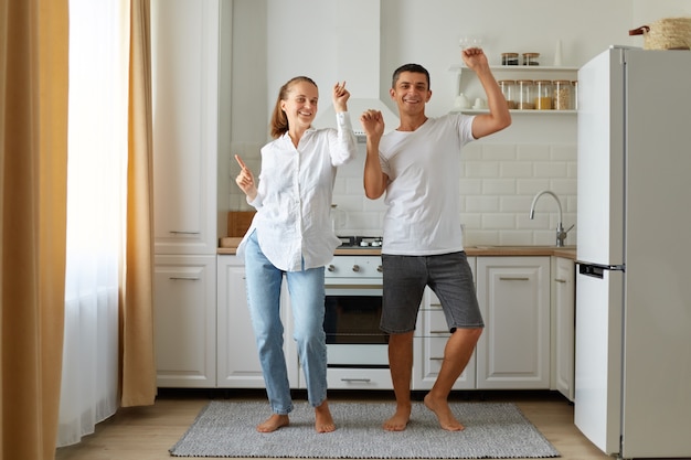 Filmación en interiores de marido y mujer positivos felices bailando, divirtiéndose juntos en la cocina, celebrando la reubicación, estando de buen humor, expresando felicidad.