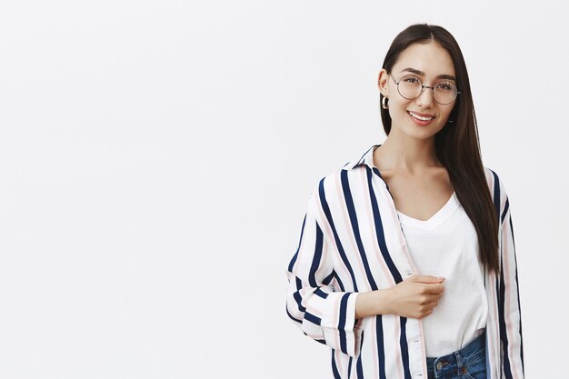 Filmación en interiores de una modelo femenina segura de sí misma con un cabello largo y hermoso y belleza natural, de pie en una moderna camisa a rayas y sonriendo ampliamente