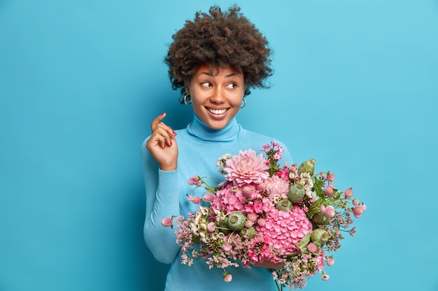 Filmación en interiores de una mujer guapa sostiene un ramo de flores mira a un lado con alegría tiene un aspecto delicado vestido con un cuello alto informal aislado sobre una pared azul