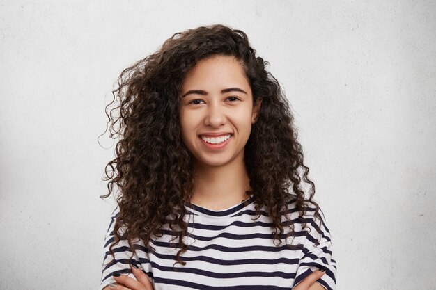 Filmación en interiores de mujer hermosa feliz con cabello oscuro y tupido y rizado, piel sana y pura, amplia sonrisa y dientes blancos uniformes