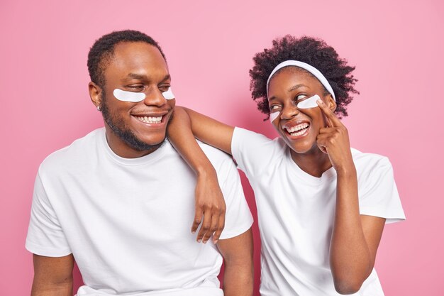 Filmación en interiores de la mujer y el hombre negros despreocupados felices miran con una sonrisa el uno al otro aplicar parches de belleza