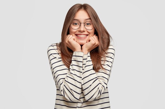 Filmación en interiores de mujer joven feliz con gafas posando contra la pared blanca