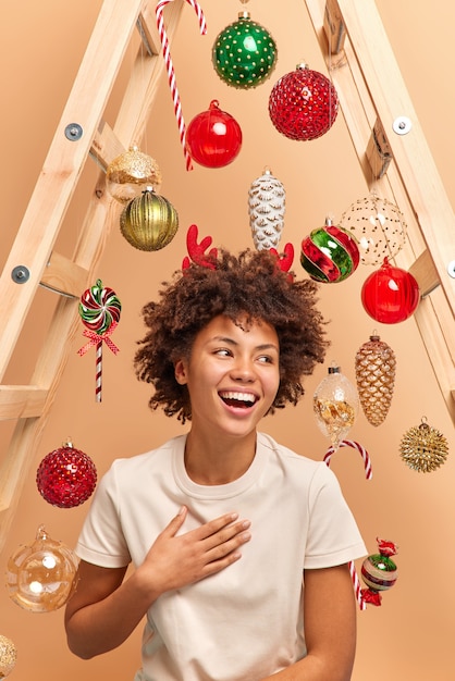 Filmación en interiores de una mujer joven muy sonriente con cabello afro rizado se ríe felizmente y mira a un lado usa astas rojas camiseta blanca casual feliz de tener vacaciones de invierno se prepara para la navidad en casa