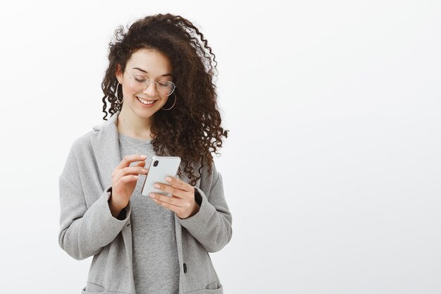 Filmación en interiores de mujer de moda feliz con aretes y gafas de moda, de pie en abrigo gris