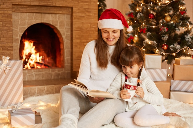 Filmación en interiores de una mujer y una niña sentada en el suelo y leyendo un libro, posando en la sala de estar decorada de forma festiva junto a la chimenea y el árbol de Navidad, Feliz año nuevo.