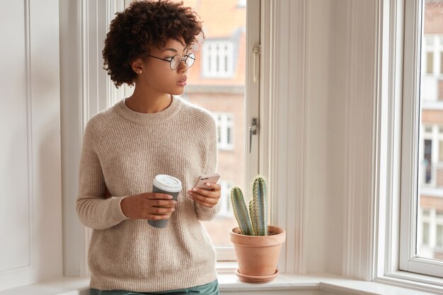 Filmación en interiores de una mujer de piel oscura pensativa utiliza un teléfono inteligente para la comunicación en línea