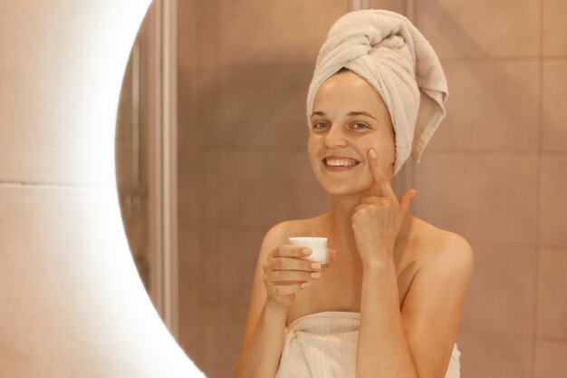 Filmación en interiores de mujer sonriente feliz de pie delante del espejo frotando crema cosmética en su rostro, poniéndose crema hidratante en su piel facial en el baño.