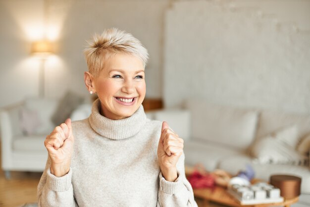 Filmación en interiores de mujeres maduras llenas de alegría de moda con suéter de cuello alto disfrutando de noticias positivas, con expresión facial extática, riendo y apretando los puños. Concepto de éxito y logros