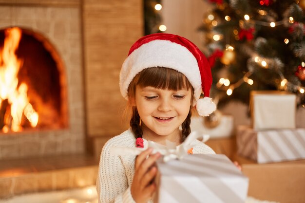 Filmación en interiores de niña feliz positiva con suéter blanco y sombrero de santa claus, sosteniendo la caja actual en las manos, posando en la sala festiva con chimenea y árbol de Navidad.