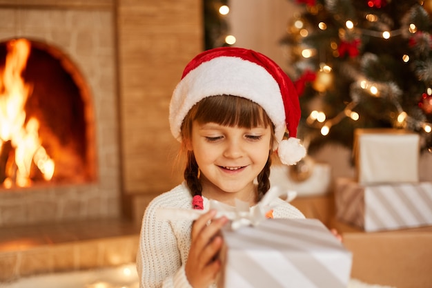 Filmación en interiores de niña feliz positiva con suéter blanco y sombrero de santa claus, sosteniendo la caja actual en las manos, posando en la sala festiva con chimenea y árbol de Navidad.