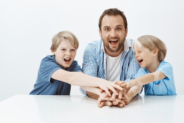 Filmación en interiores de niños atractivos alegres y padre sentado a la mesa, tomados de la mano, riendo y gritando de felicidad. papá haciendo equipo con hijos para hacer un regalo para mamá