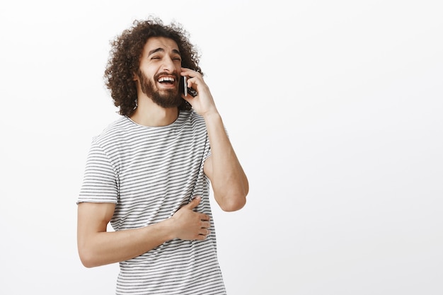 Foto gratuita filmación en interiores de un novio atractivo alegre con barba y peinado afro, mirando a un lado mientras habla por teléfono inteligente, riendo a carcajadas