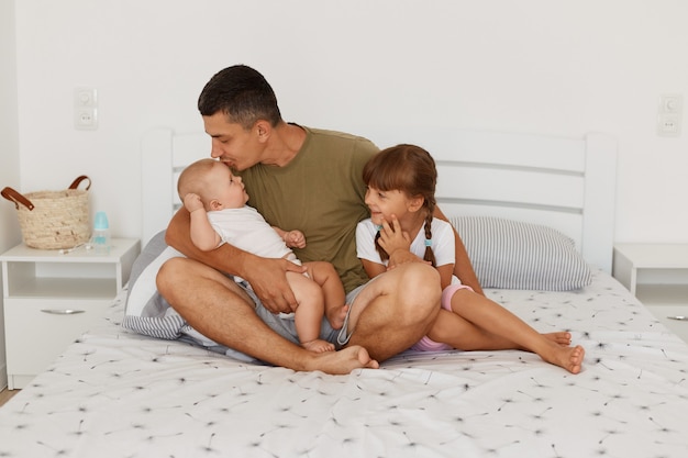 Filmación en interiores de un padre feliz posando con sus hijos pequeños, besando a su bebé, un hombre atractivo morena con ropa de estilo casual que pasa tiempo con sus hijos.